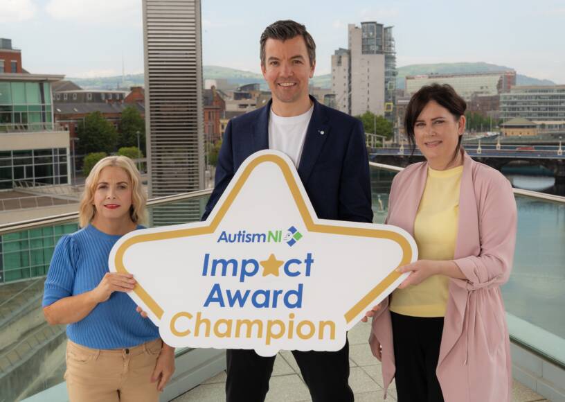 Three people standing in front of windows with the River Lagan in the Background, holding an Autism NI Impact Award logo board.