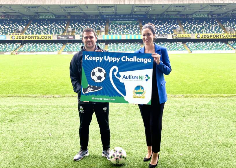 Two people in a football field smile and hold a poster that reads 'Keepy Uppy Challenge'