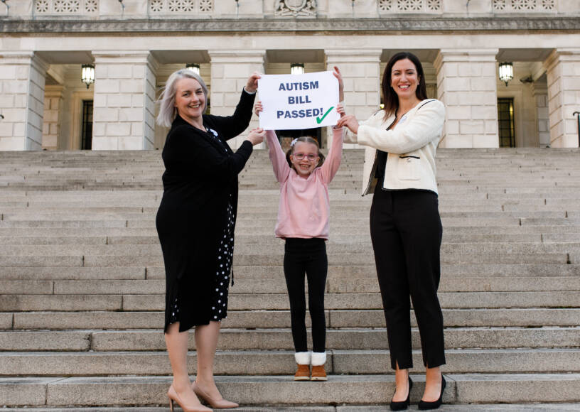 Autism NI members and young child holding up a sign that reads 'Autism Bill passed!'