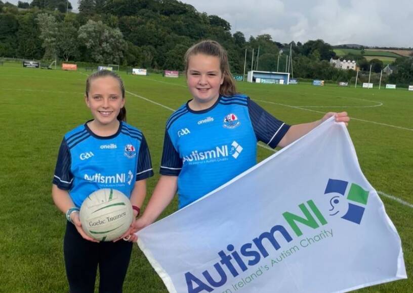 Two children standing on a pitch with a football, holding up an Autism NI flag