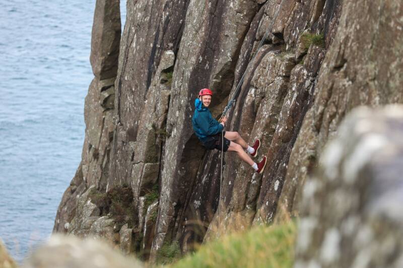A man is wearing a harness and has descended halfway down a cliff.