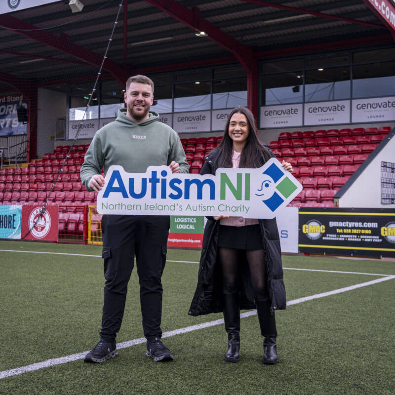 Two people standing on a football pitch holding an Autism NI logo board in support of Northern Ireland's autism charity.