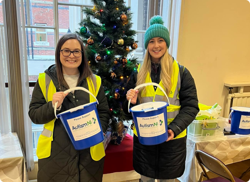 Autism NI volunteers are fundraising and making themselves seen by wearing high-vis jackets and holding collection buckets in front of a Christmas tree.