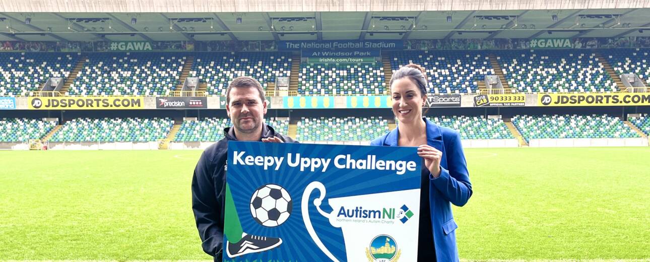 Two people in a football field smile and hold a poster that reads 'Keepy Uppy Challenge'