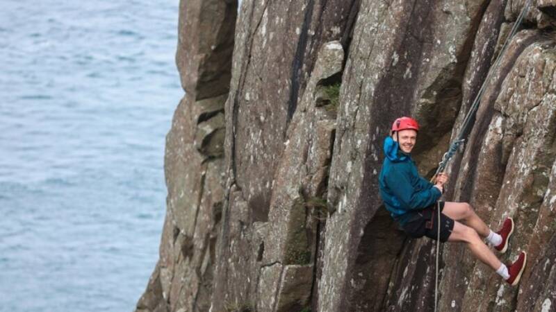 A man is wearing a harness and is halfway down a cliff looking side on smiling.