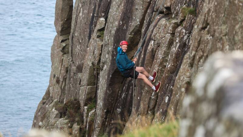 A man is wearing a harness and has descended halfway down a cliff.
