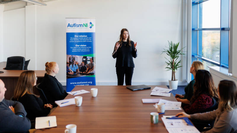 An autism trainer stands at the top of the room and speaks to the delegates who are all sitting around a rectangular table looking at her.