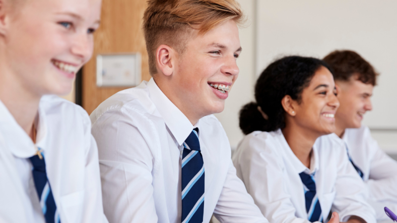 A row of four students in uniform sitting smiling while listening in class.