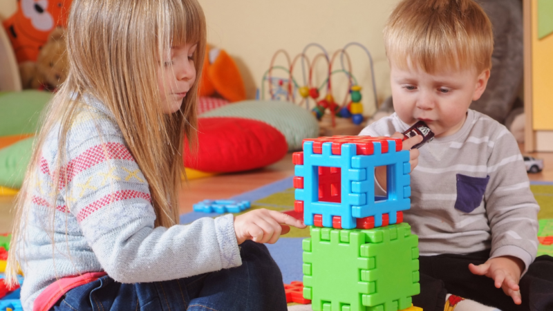 Two young children sitting on the floor, playing together - building a tower of colourful blocks.