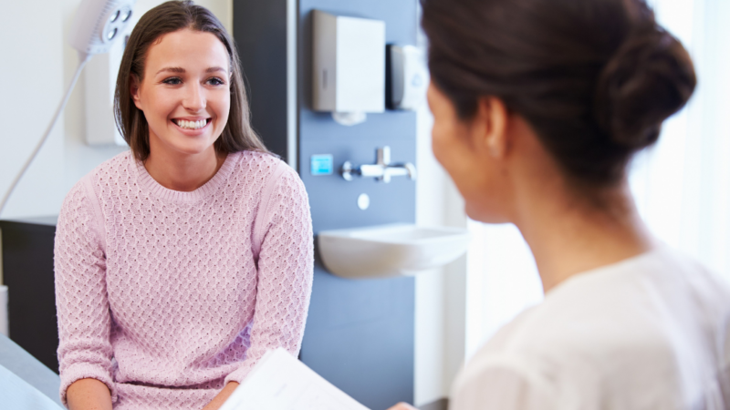 A woman is sitting on a hospital bed talking to a practitioner in the foreground of the photo.