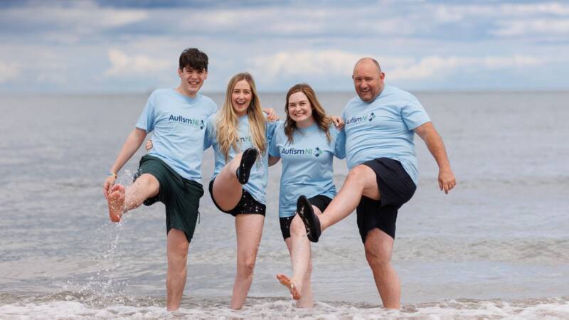 Four adults stand in the shallow waters kicking one leg up and smiling. They are all wearing Autism NI t-shirts and promoting a Sea Dip fundraising challenge for Autism NI.