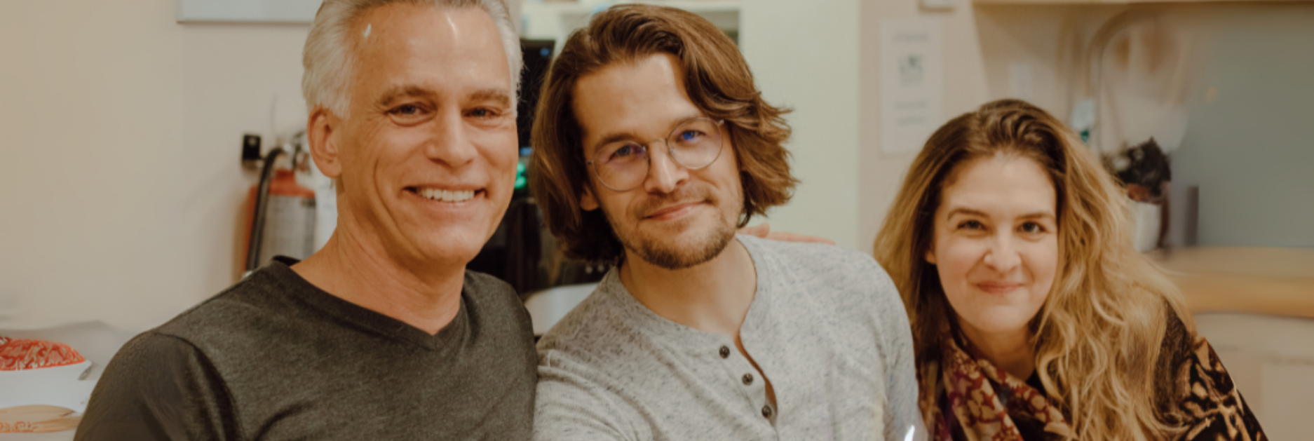 3 adults sitting beside each other smiling in a kitchen area