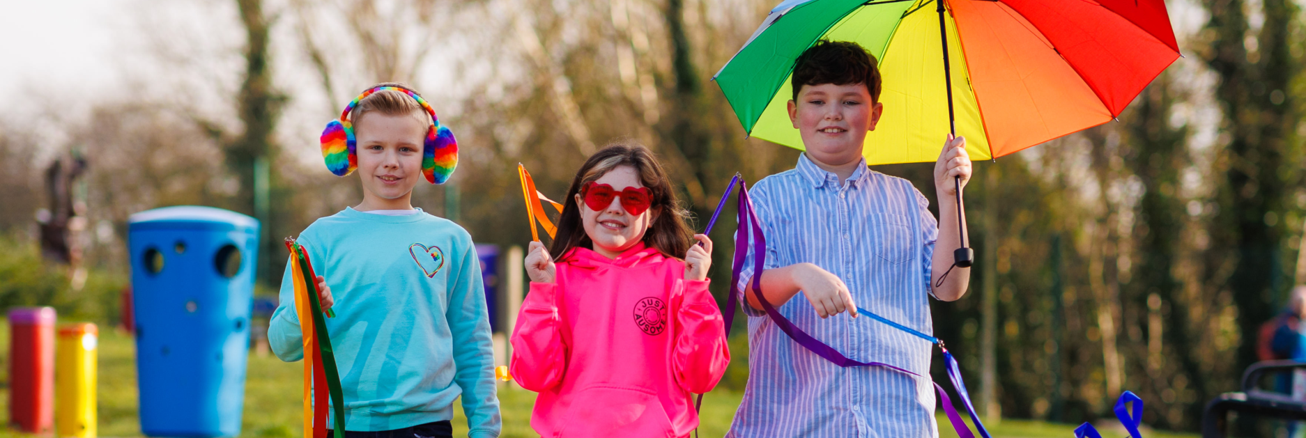 Three children in a park, wearing and holding colourful items, and walking towards the camera