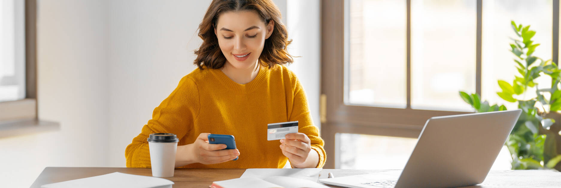 A woman sitting in front of her laptop and typing on her phone, with her credit card in her other hand