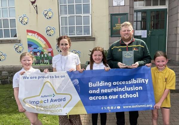 A teacher and pupils stand outside a school holding a banner that reads 'Autism NI Impact Award Educations Champion', after receiving autism training for education.