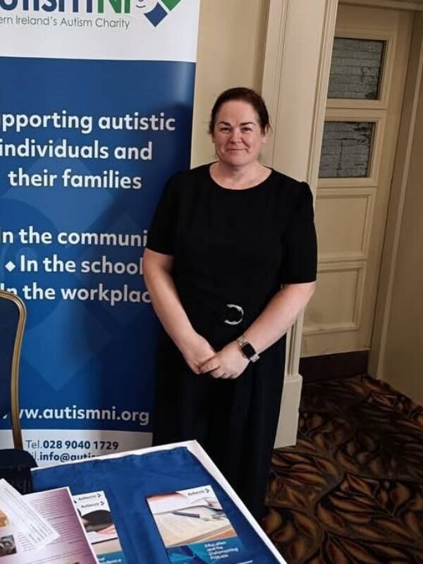 A person is standing beside an Autism NI pop-up stand and behind a table with Autism NI resources on it.