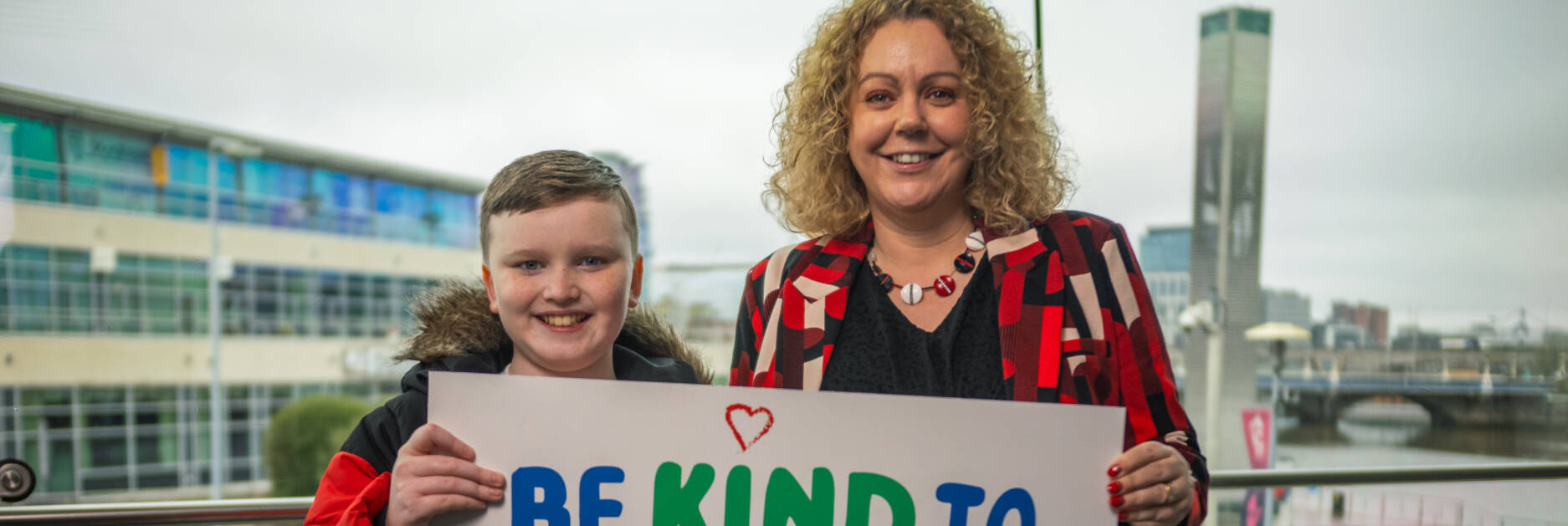 A woman and child smile and holding a sign reading 'Be kind to different minds' for autism awareness and acceptance.