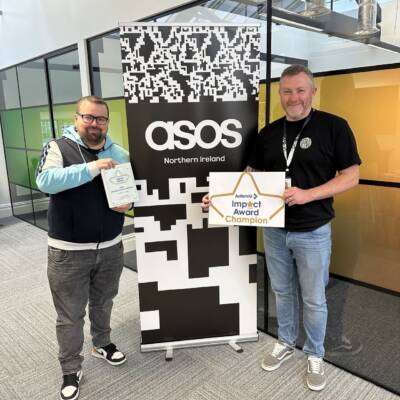 Two members of ASOS smile in front of an ASOS poster and hold their Autism NI impact award