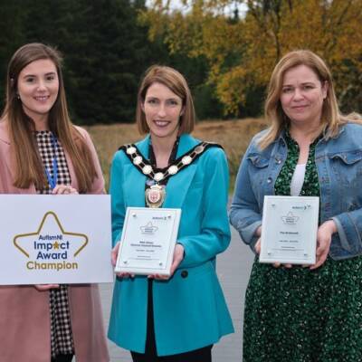 Three women smile and pose together outdoors holding Autism NI impact awards