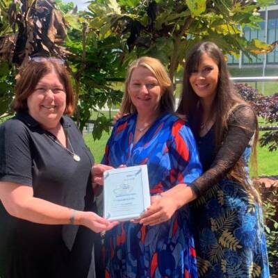 A group of women smile and pose outdoors with an impact award