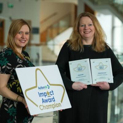 Two women smile and pose with impact awards and an impact award champion poster