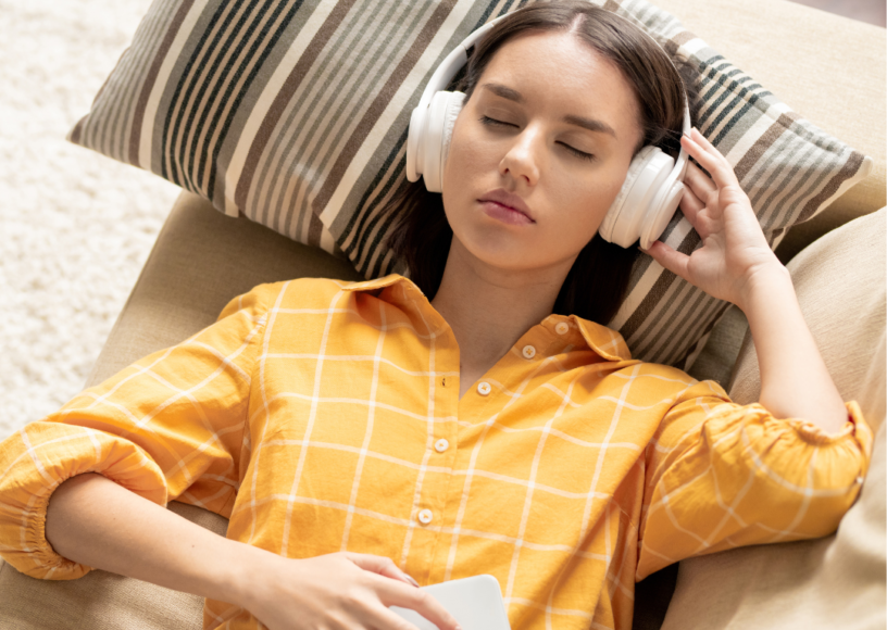 An adult female lying on a sofa with headphones, eyes closed, relaxing.